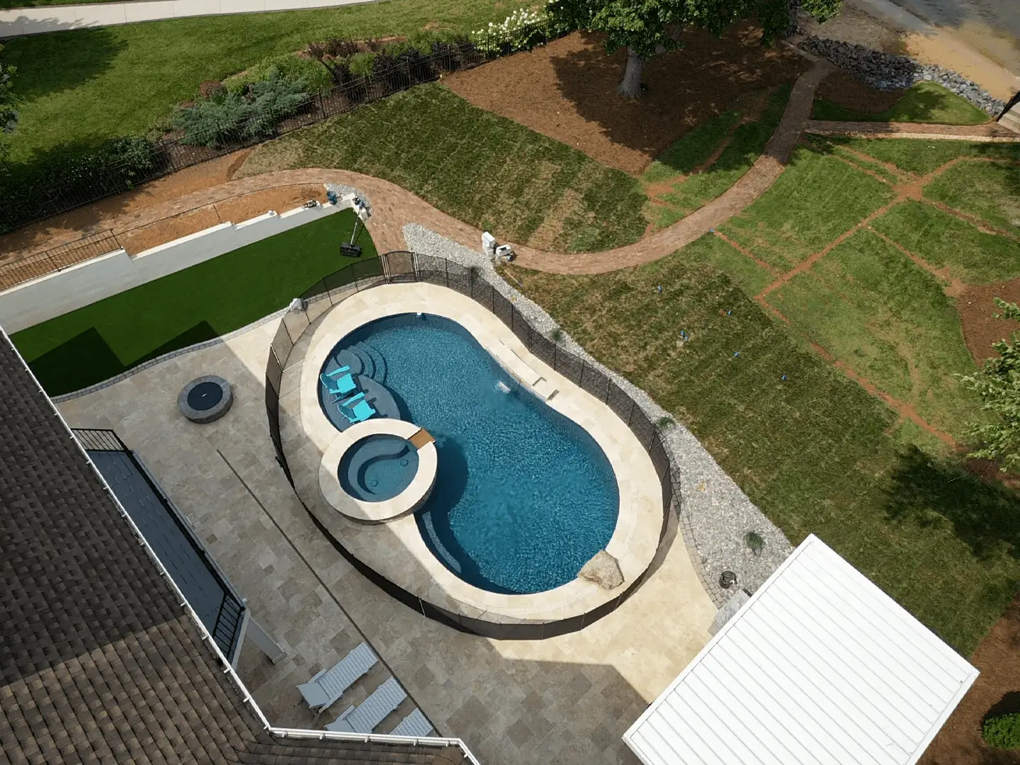 Aerial view of a backyard with an oval swimming pool and attached hot tub, surrounded by a stone deck. There's a lawn chair, a patch of artificial grass, and pathways on a newly landscaped lawn. A person is walking near the pool.