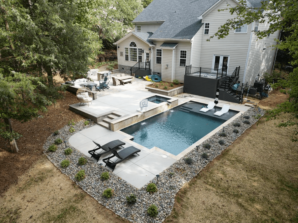 Aerial view of a modern backyard featuring a rectangular swimming pool with lounge chairs, a basketball hoop, and surrounding patio furniture. The area is landscaped with small shrubs and bordered by trees, adjacent to a gray, two-story house.