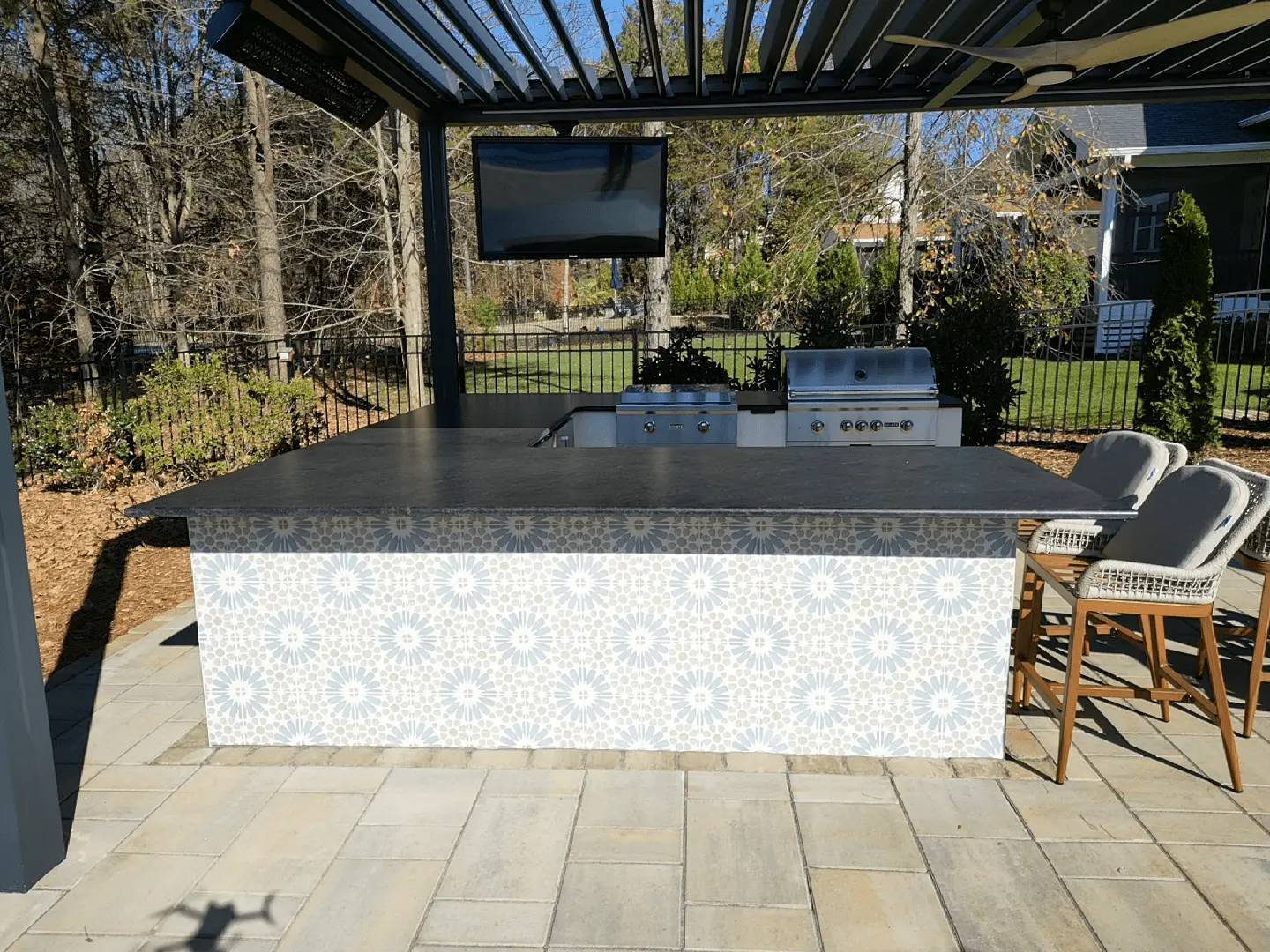 Outdoor kitchen with a patterned tile island featuring two wicker chairs, a built-in grill, and a TV mounted under a pergola. Trees and shrubs are visible in the background, surrounded by a fence.