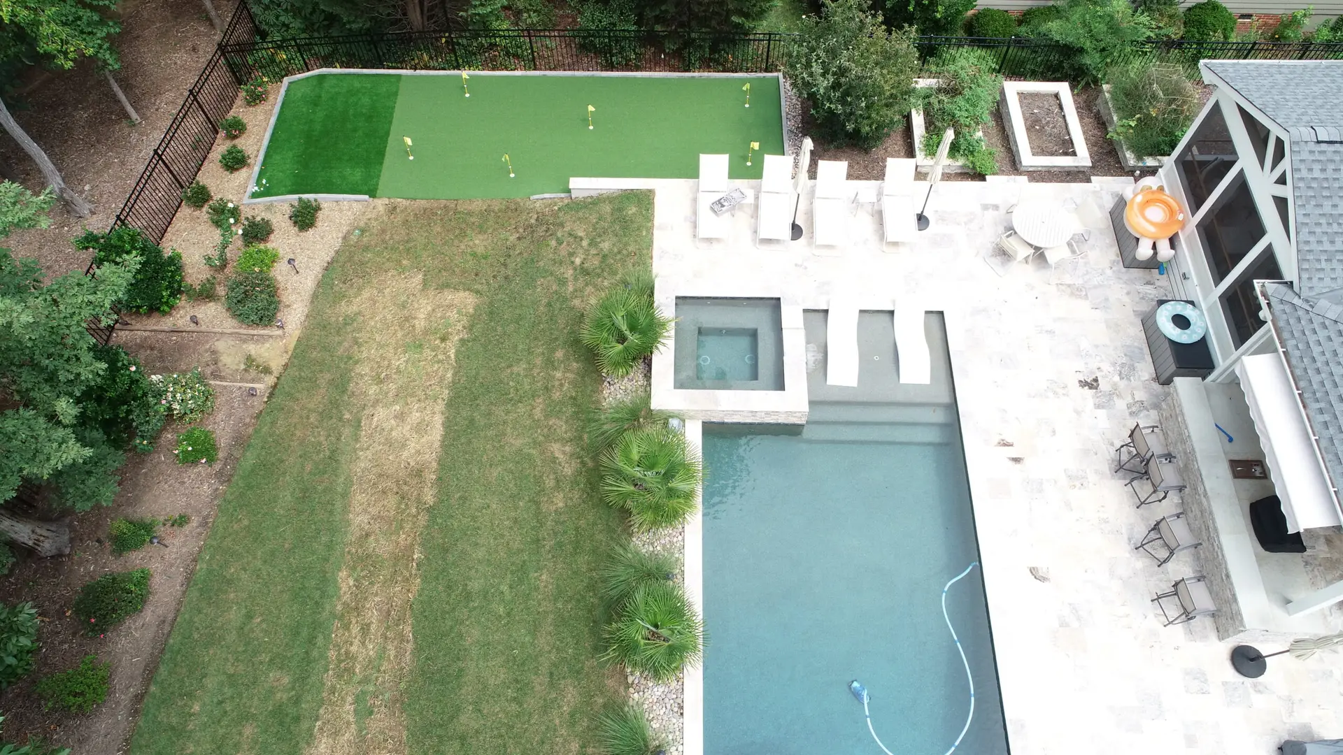 Aerial view of a backyard featuring a rectangular pool with a hot tub, surrounded by a stone patio with chairs. Adjacent is a putting green and landscaped areas with trees and shrubs.