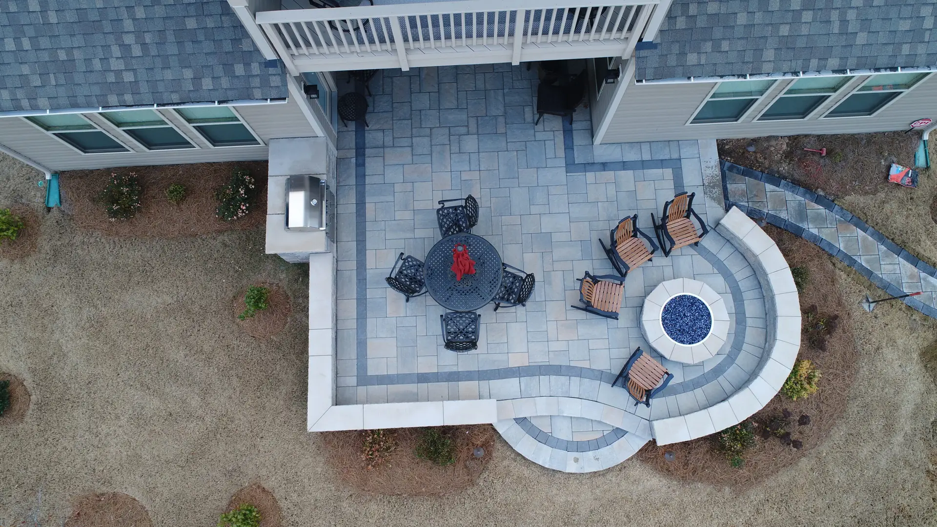 Aerial view of a backyard patio with a circular fire pit surrounded by four chairs, a round table with four chairs, and a gas grill. The patio is paved with stone tiles and bordered by a garden. A paved path curves around the patio area.