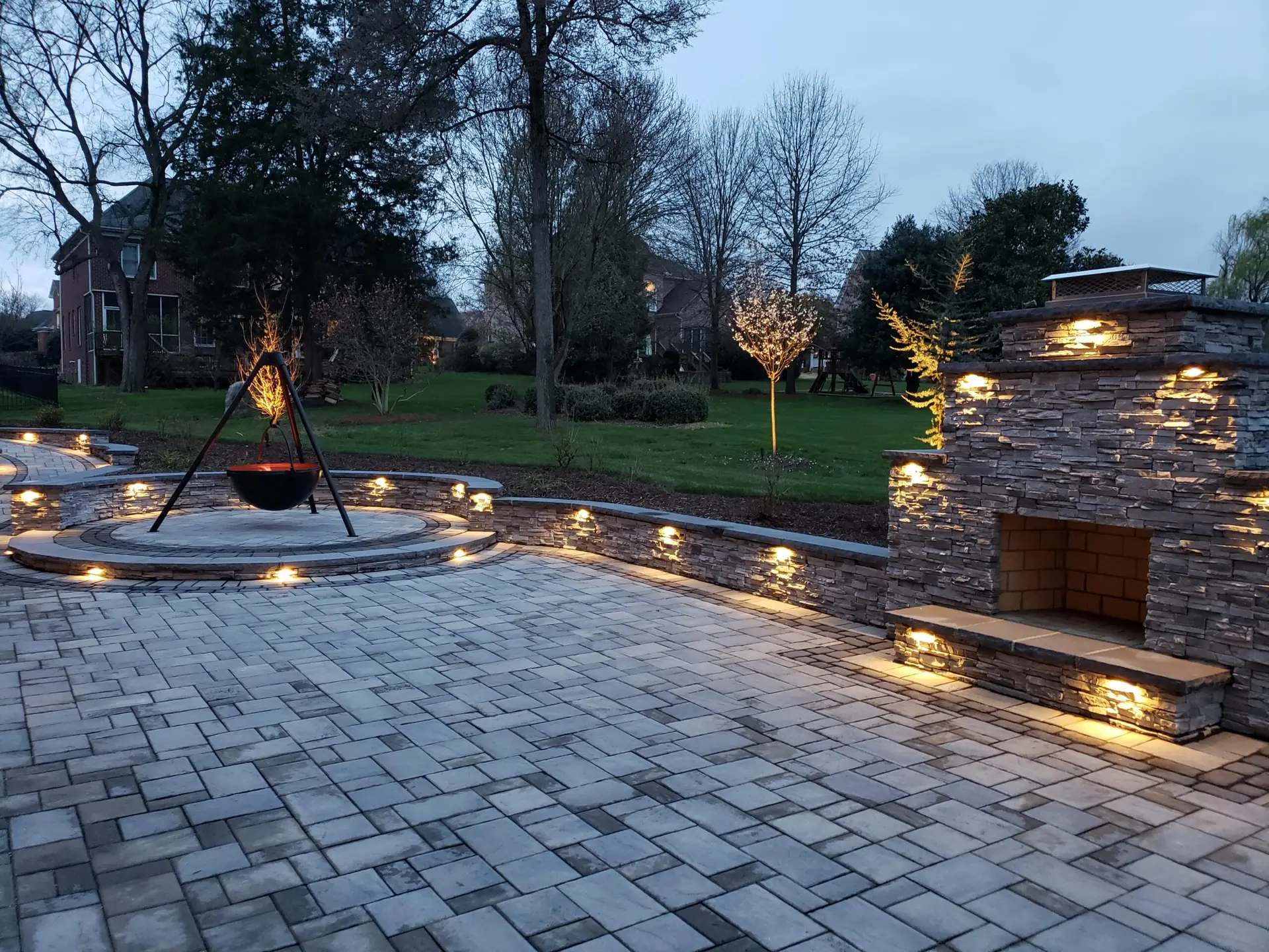 A spacious backyard patio features a stone fireplace and a central circular fire pit, both illuminated by soft, warm lights. The paved area is surrounded by trees and lush greenery, creating a cozy and inviting outdoor space.
