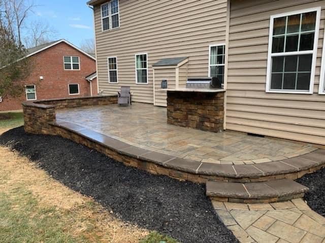 A modern backyard patio with beige siding and stone accents features a built-in grill and a chair. The patio is elevated with stone steps and surrounded by a mulch border. Nearby, a red brick house and trees are visible in the background.