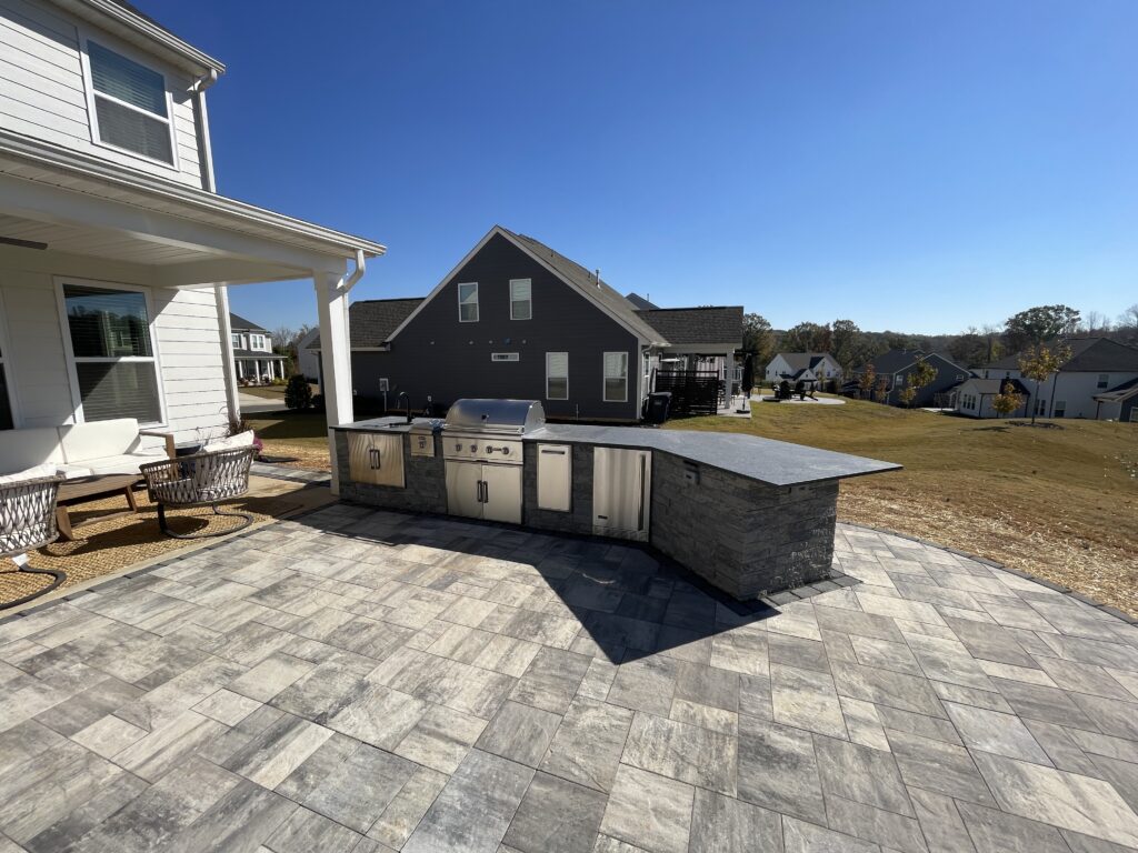 Outdoor patio with stone-tiled flooring features a built-in stainless steel grill and counter. To the left, white patio chairs sit near a house, while in the background, suburban homes are visible under a clear blue sky.