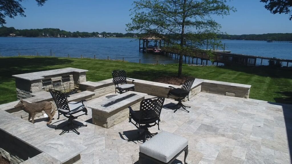 A serene lakeside patio featuring stone seating and black metal chairs surrounding a rectangular fire pit. A tree provides shade, and a wooden dock extends into the tranquil water, with a clear blue sky above.