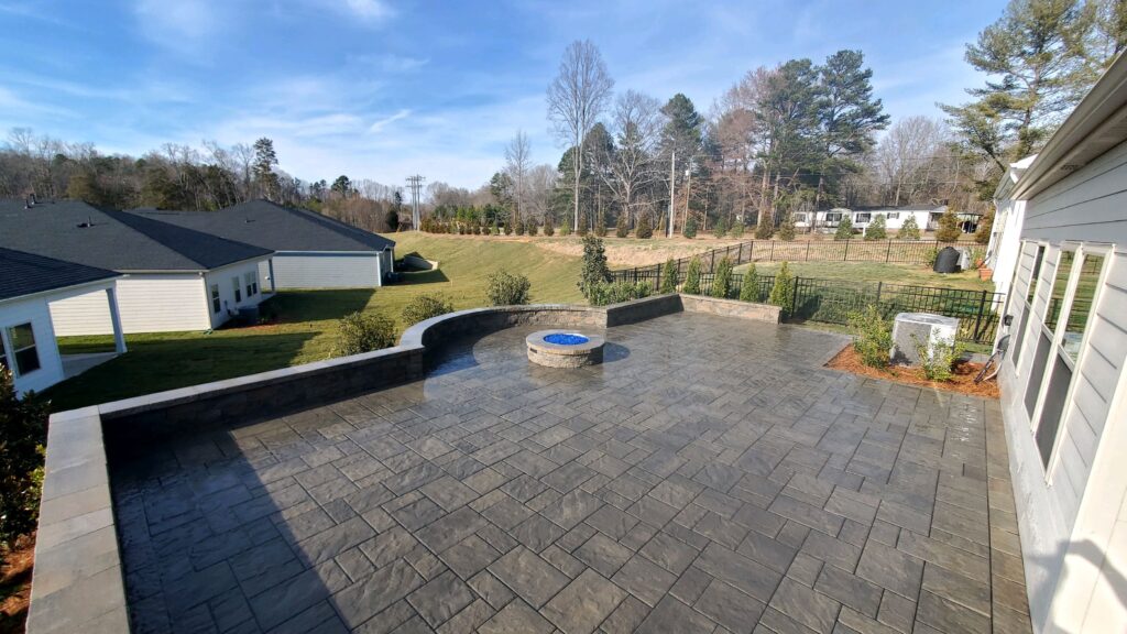 Elevated patio with stone flooring and a circular fire pit in the center. It overlooks a grassy lawn and neighboring houses, surrounded by trees under a clear blue sky.