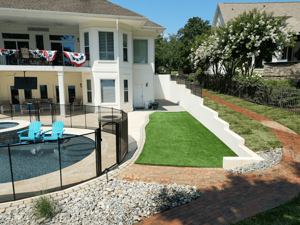 Backyard with a circular pool surrounded by a safety fence, two blue Adirondack chairs, and a small artificial grass area. A brick path leads to a two-story house decorated with red, white, and blue banners. Trees and bushes line the property.