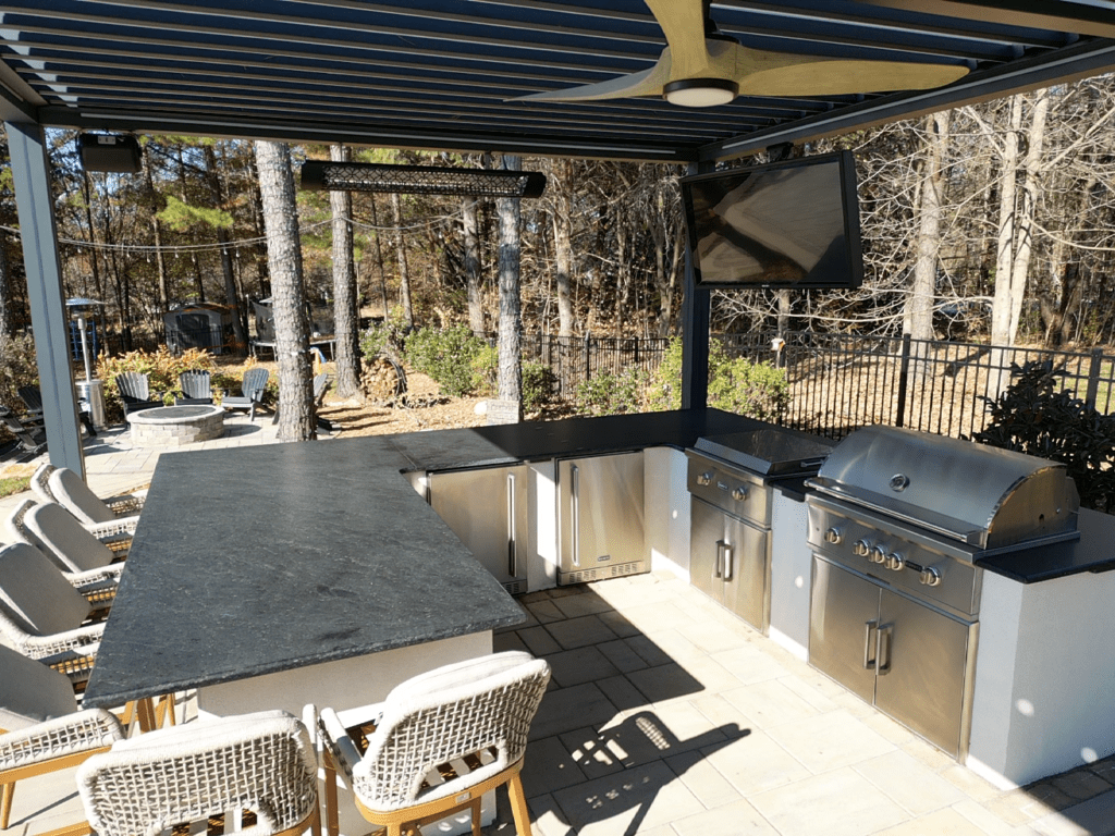 Outdoor kitchen with stainless steel appliances, including a grill and fridge, under a pergola. There's a large black countertop with bar stools. A TV is mounted above the countertop, and a fire pit is visible in the background amidst trees.