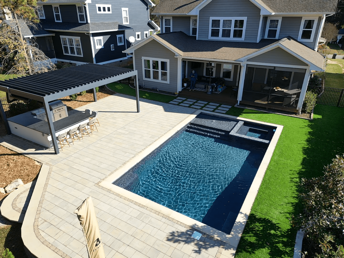 Aerial view of a backyard with a rectangular swimming pool surrounded by a patio. Adjacent to the pool is a pergola-covered outdoor kitchen with bar seating. The area is landscaped with green grass and bordered by houses.