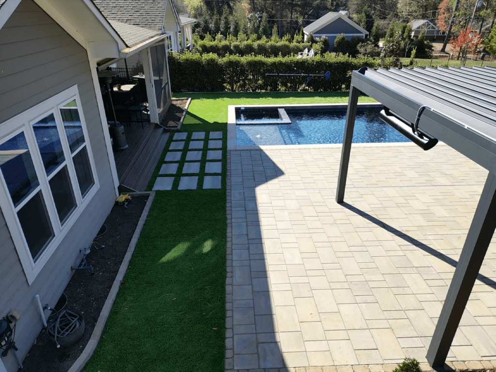 A backyard with a swimming pool and hot tub features a large paved patio area. Adjacent is an artificial grass patch with stepping stones leading to a wooden deck connected to a gray house. A pergola provides partial shade.