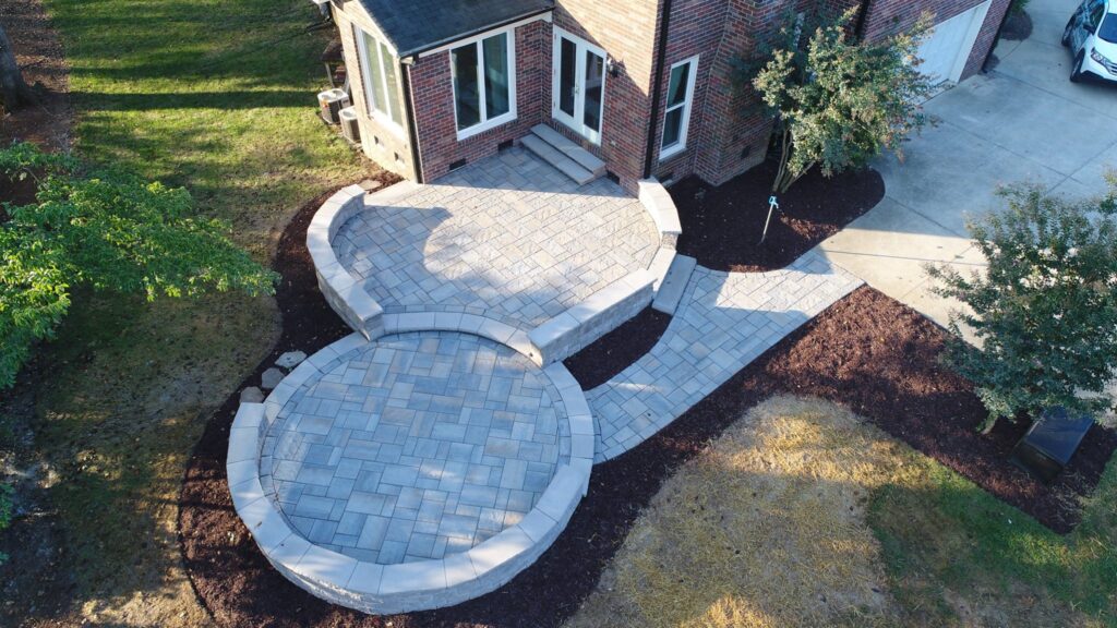 Aerial view of a landscaped patio with grey stone pavers outside a brick house. The patio has a circular section and rectangular steps leading to the entrance. The area is surrounded by mulch and small trees.