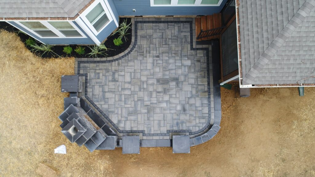 Aerial view of a newly constructed outdoor patio with gray stone pavers in a geometric pattern. The patio is adjacent to a blue house and features a built-in corner seating area and steps leading to the entrance. Surrounding ground is dry and grassy.