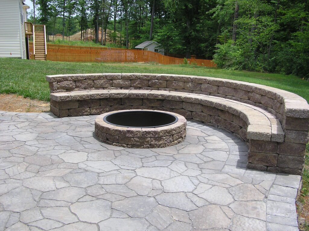 Curved stone bench seating surrounds a circular fire pit on a paved patio. The area is bordered by a lush green lawn, with trees and a wooden fence in the background.