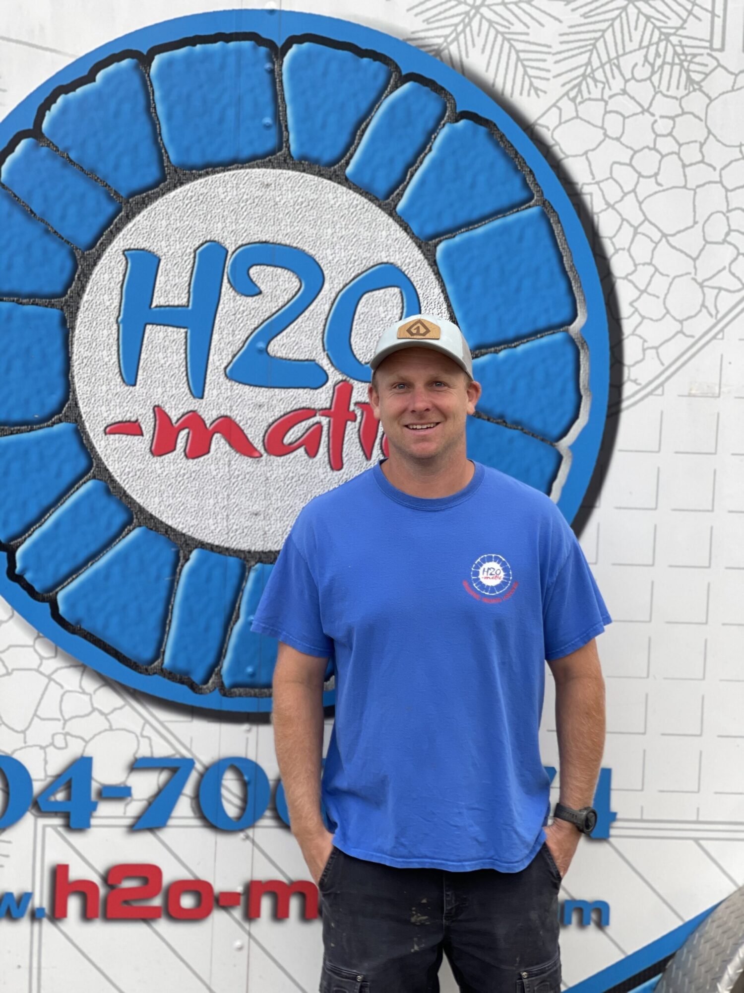 Man in a blue T-shirt and a light cap stands smiling in front of a promotional sign for H2O-matic, featuring a large water-themed graphic. He's positioned slightly to the side, hands in pockets.