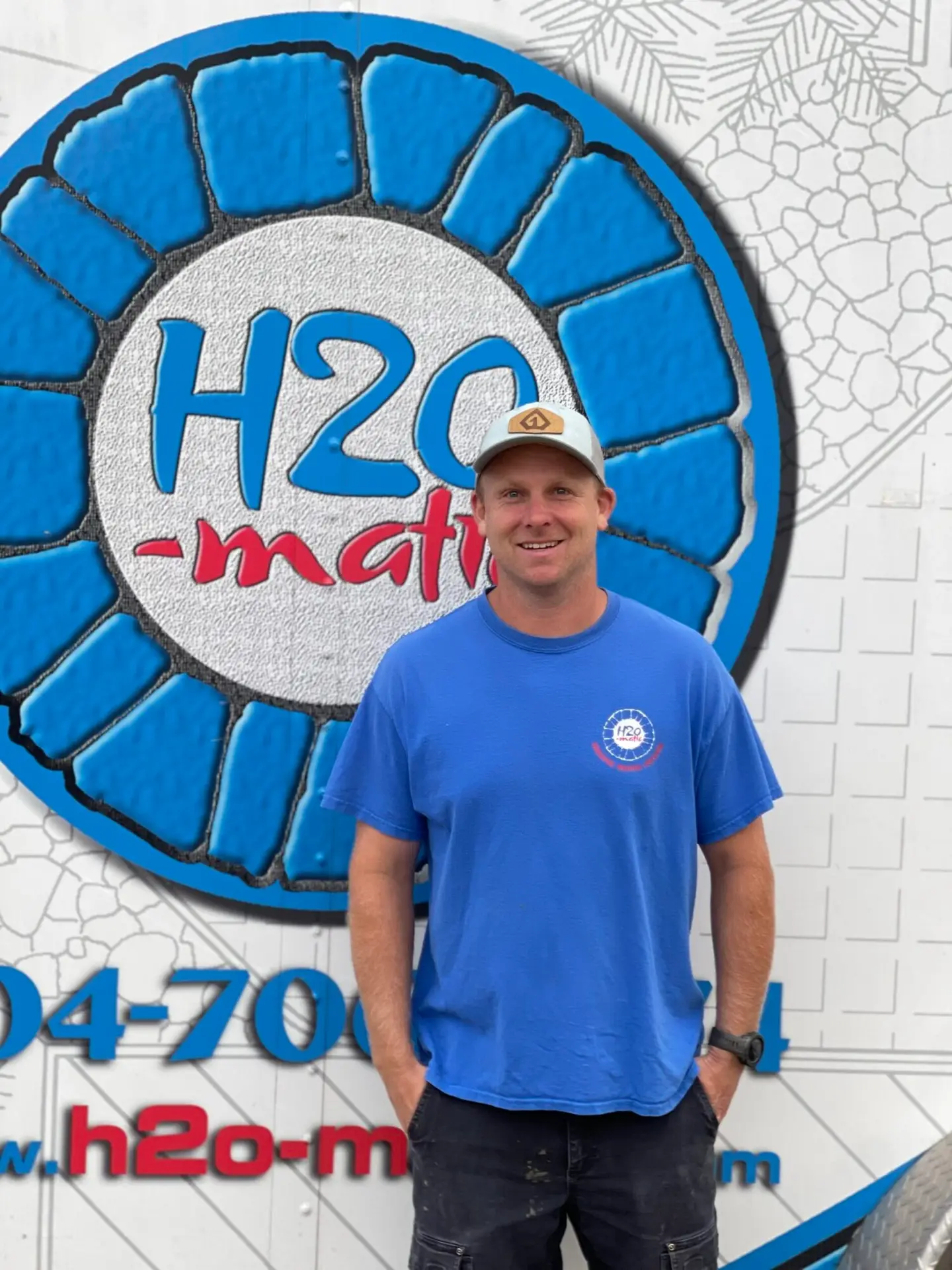 Man in a blue T-shirt and a light cap stands smiling in front of a promotional sign for H2O-matic, featuring a large water-themed graphic. He's positioned slightly to the side, hands in pockets.