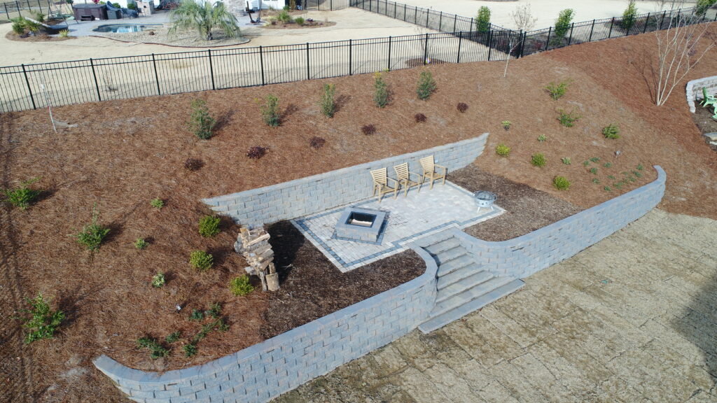 Aerial view of a landscaped hillside with two wooden chairs on a brick patio. The area features a small pyramid-shaped stone structure and a metal fire pit. Young plants are spread across the sloped, mulched ground, with a black metal fence at the top.