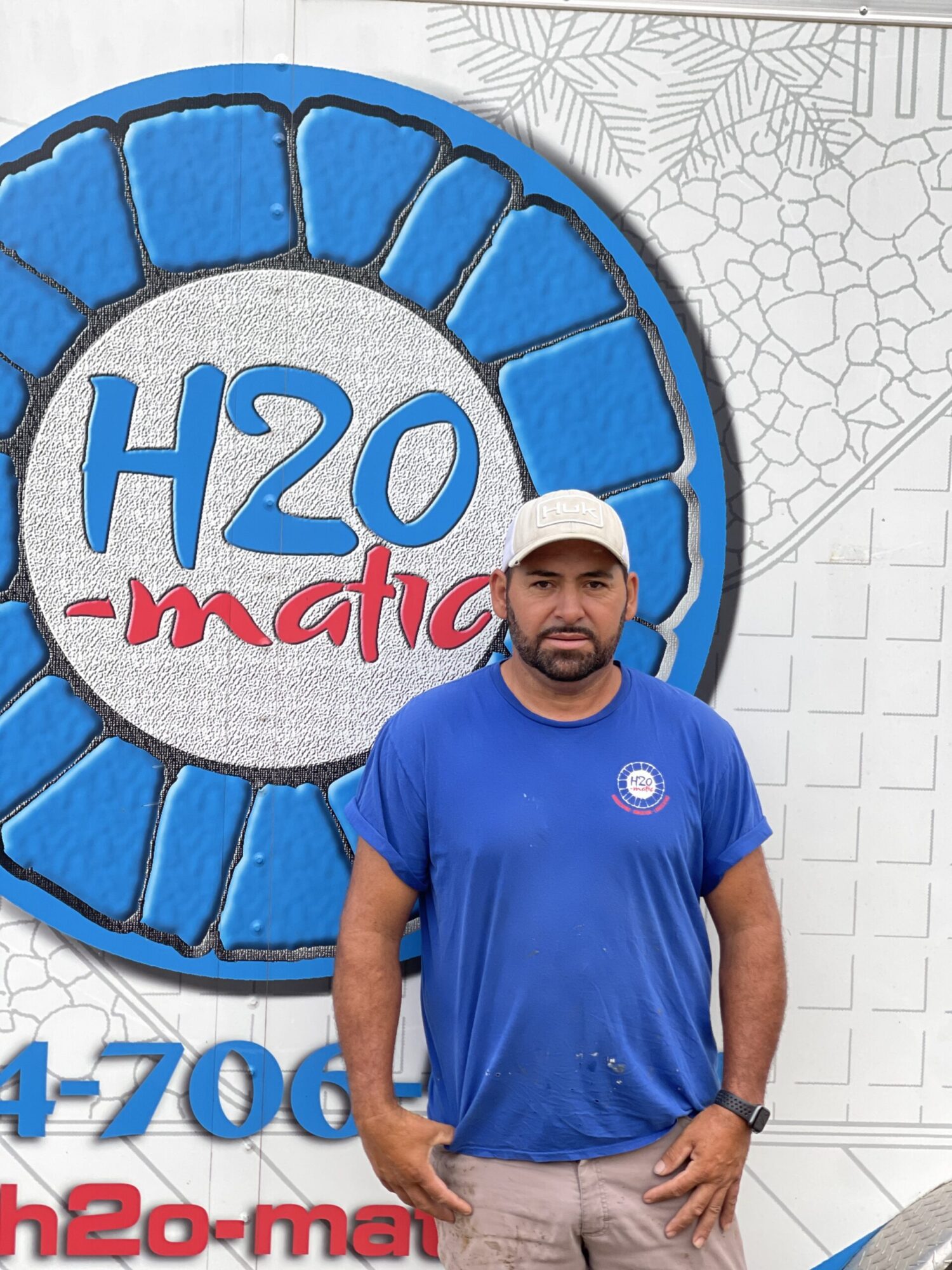 A man in a blue t-shirt and beige shorts stands in front of a vehicle with a large, circular blue logo that says "H2O-matic." He wears a baseball cap and has a serious expression. Parts of a phone number are visible on the vehicle.