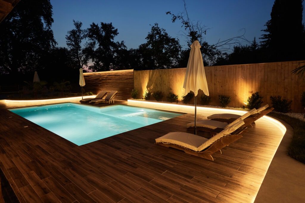 A modern backyard pool illuminated at night. The pool area features wooden decking, two cushioned lounge chairs with umbrellas, and ambient lighting along the perimeter. Tall trees and a wooden fence surround the serene setting.