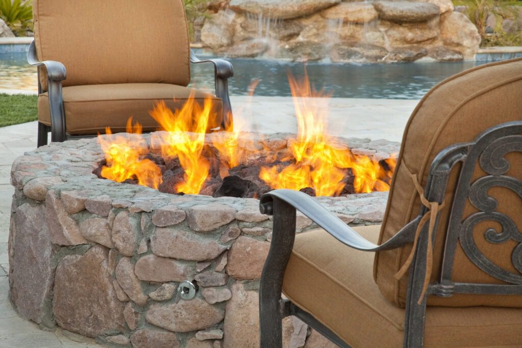 A stone fire pit with bright orange flames is surrounded by two cushioned chairs. In the background, there's a swimming pool and natural rock formations.