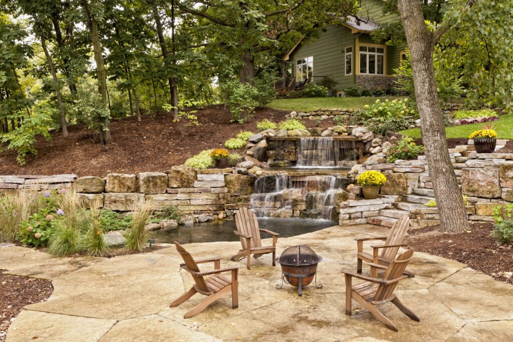 A serene backyard with a flowing waterfall cascading over rocks into a small pond, surrounded by lush greenery. Four wooden chairs encircle a small fire pit on a stone patio, with a cozy house in the background.