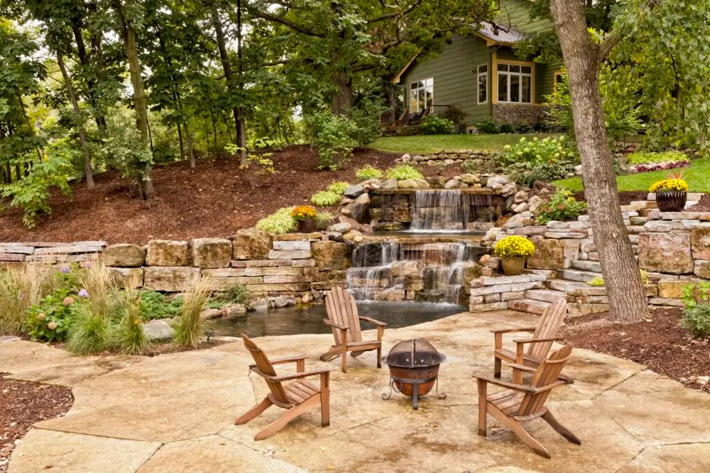 A serene backyard with a flowing waterfall cascading over rocks into a small pond, surrounded by lush greenery. Four wooden chairs encircle a small fire pit on a stone patio, with a cozy house in the background.