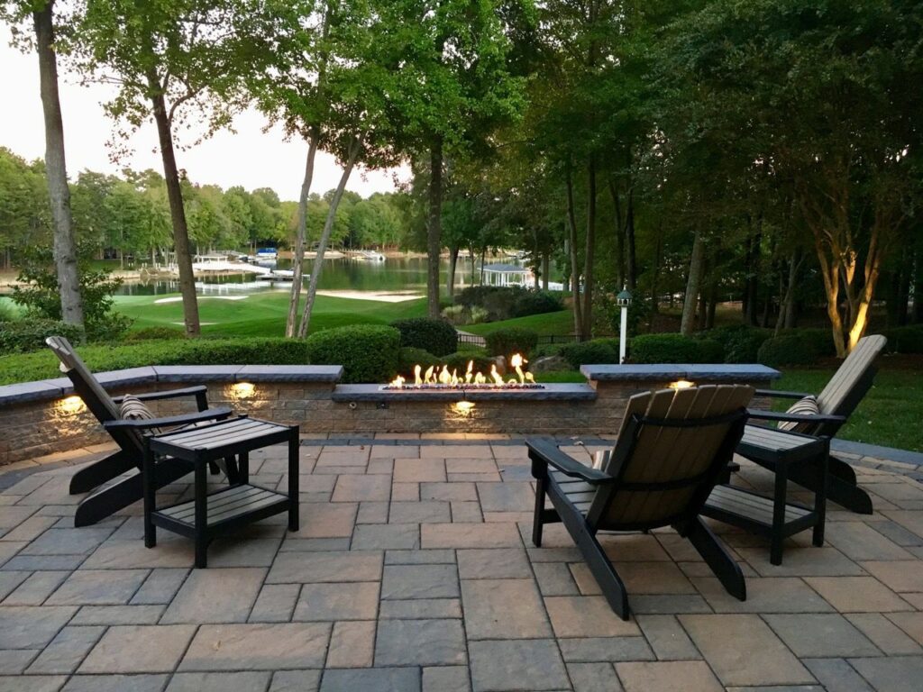 A serene backyard patio with four wooden chairs around a rectangular fire pit. Beyond, a lush lawn and trees frame a tranquil lake view, creating a peaceful outdoor setting.