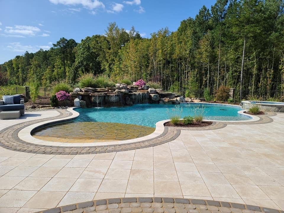 A luxurious backyard pool with a natural stone waterfall feature. The pool is surrounded by a tiled deck and bordered by lush greenery and trees. A seating area is visible in the background under a blue sky.