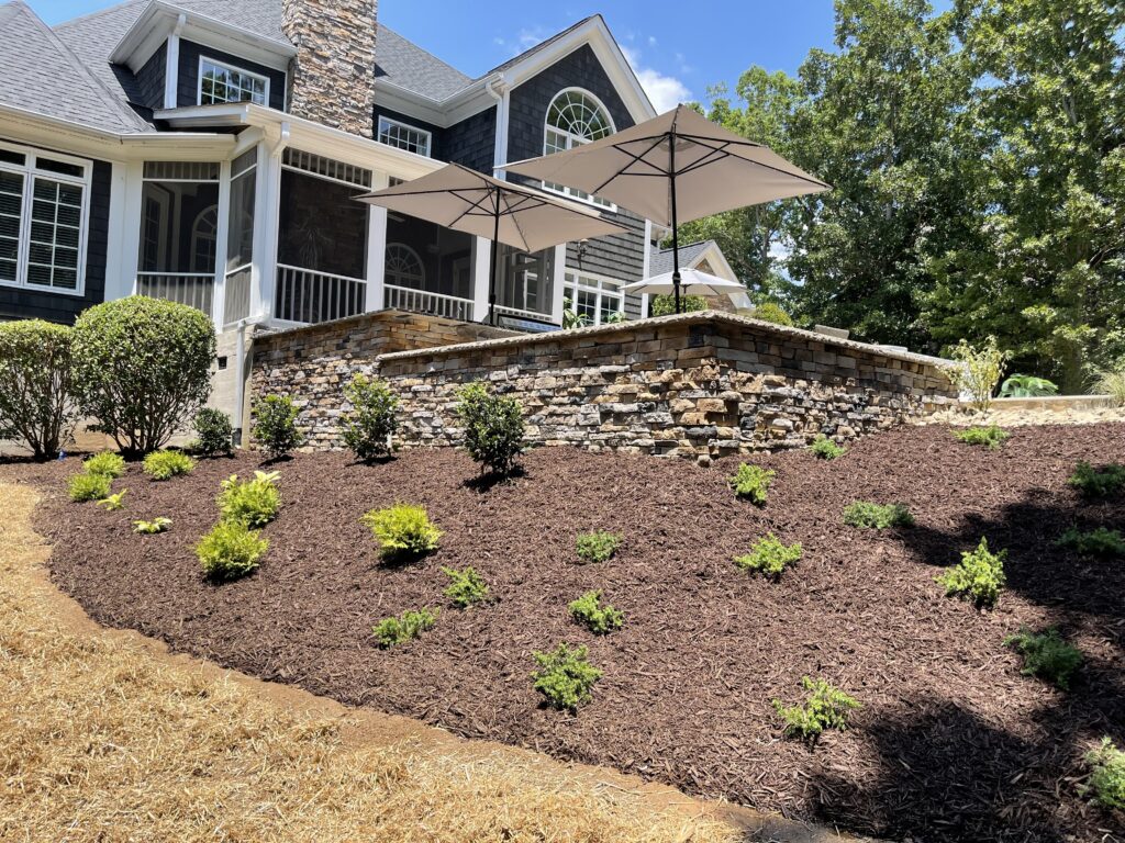 A landscaped backyard with a stone retaining wall, mulched flower beds, and small green plants. A house with a screened porch and two large umbrellas on a patio is visible in the background.