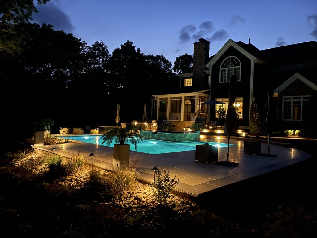 A luxurious house with a warmly lit swimming pool at dusk. The pool area is surrounded by trees and landscaped plants, creating a serene ambience. The glow from the pool and house lights illuminates the scene against a darkening sky.