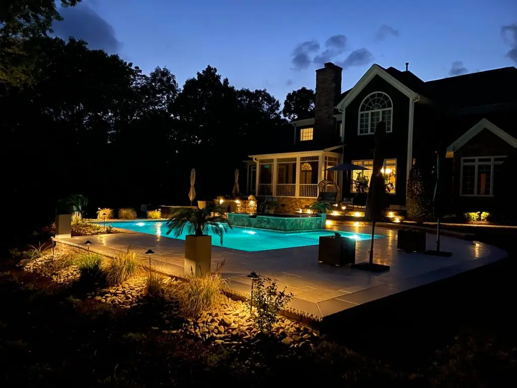 A luxurious house with a warmly lit swimming pool at dusk. The pool area is surrounded by trees and landscaped plants, creating a serene ambience. The glow from the pool and house lights illuminates the scene against a darkening sky.