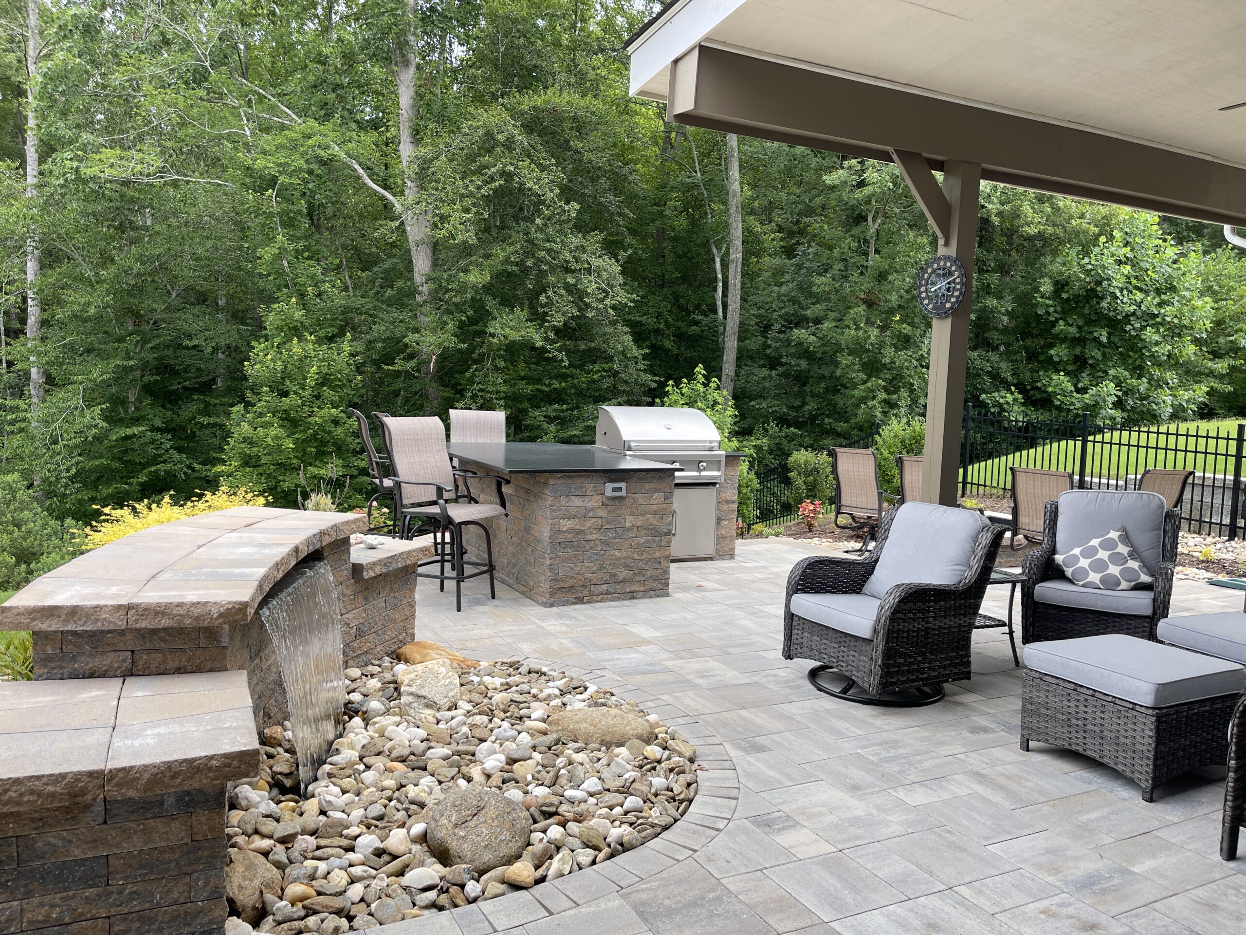 A spacious outdoor patio featuring cushioned chairs, a stone bar, a grill, and a dining area. The patio is surrounded by a lush green forest, and the ground is paved with stone tiles with a pebble area. A wall clock hangs nearby.