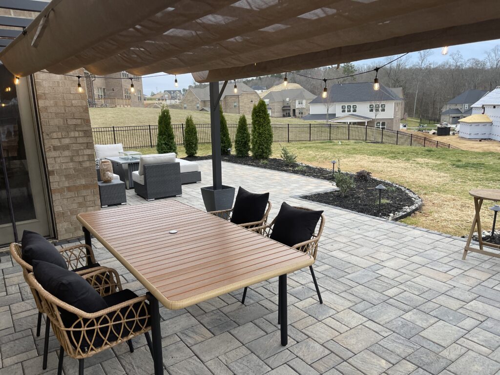 A backyard patio features a wooden table with black cushioned chairs under a canopy. Beyond, there are patio sofas, a small garden plot, and views of neighboring houses and trees.