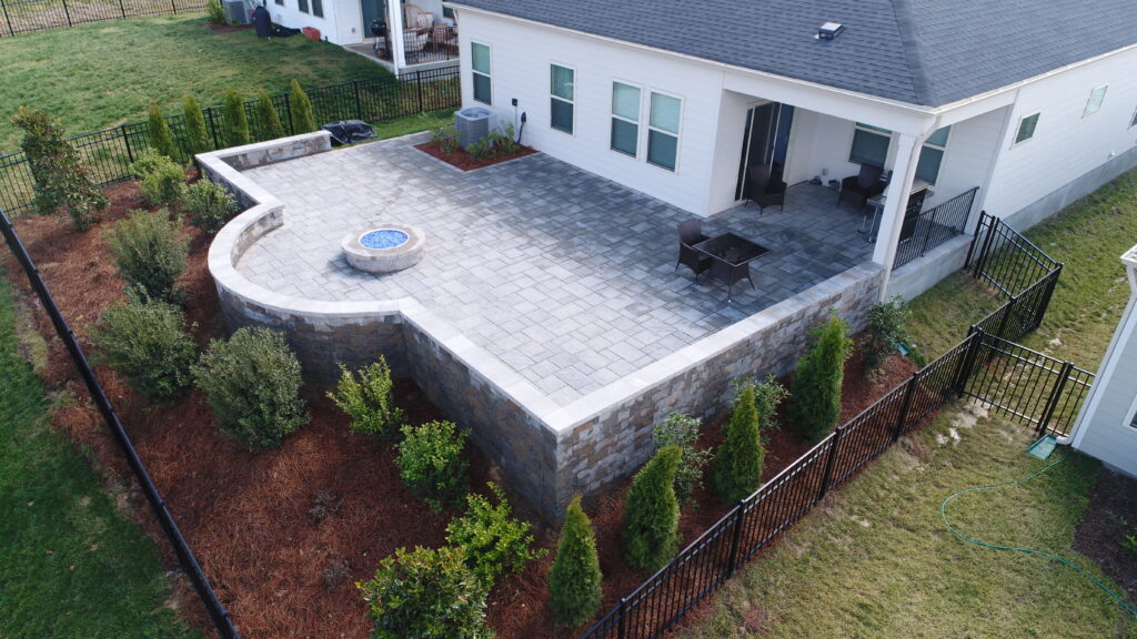 Aerial view of a backyard with a stone patio featuring a fire pit, table, and chairs. The patio is elevated with a retaining wall, surrounded by neatly planted shrubs and trees. A black fence encloses the yard.