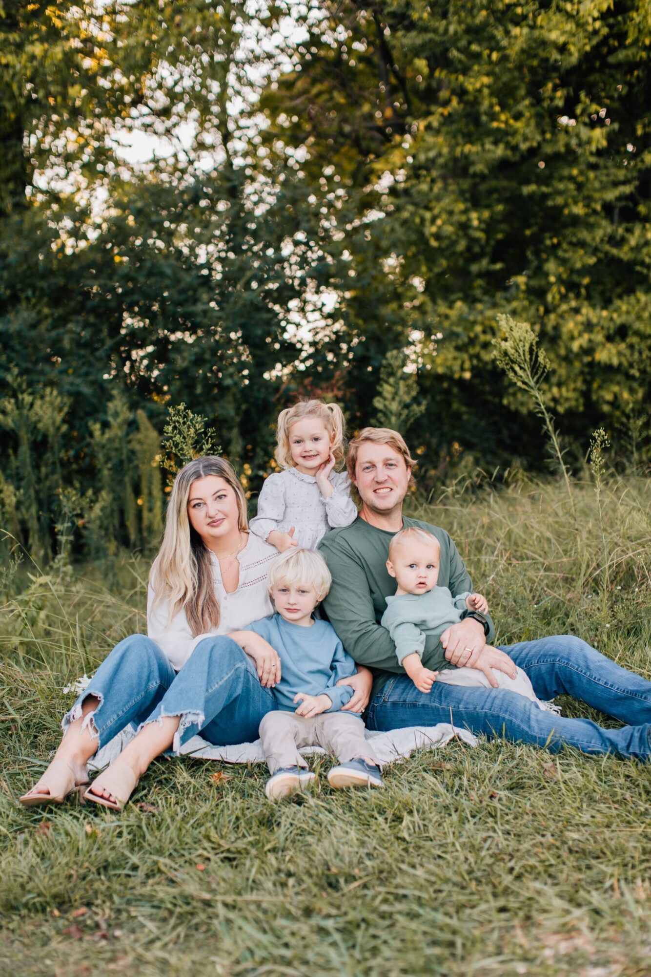 A family of five sits on the grass in a park. The parents, wearing casual clothes, hold three young children. The children, two boys and a girl, smile at the camera. Trees and greenery form the background.