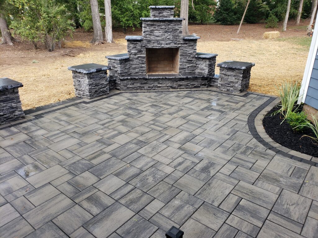 Outdoor patio with a gray stone fireplace and matching stone pillars. The ground is paved with large gray tiles in a geometric pattern. Trees and mulch are in the background.