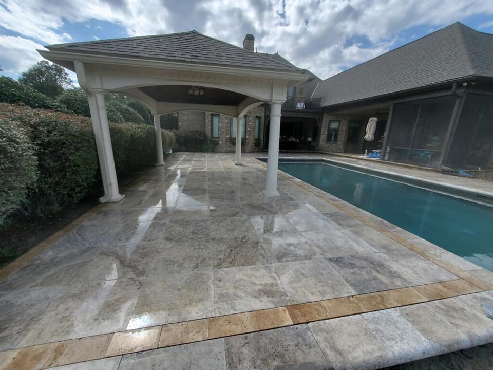 A backyard with a glossy stone patio and a rectangular swimming pool. A covered seating area with a roof and pillars is on the left. The sky is partly cloudy, and hedges border the patio. A house with large windows is in the background.