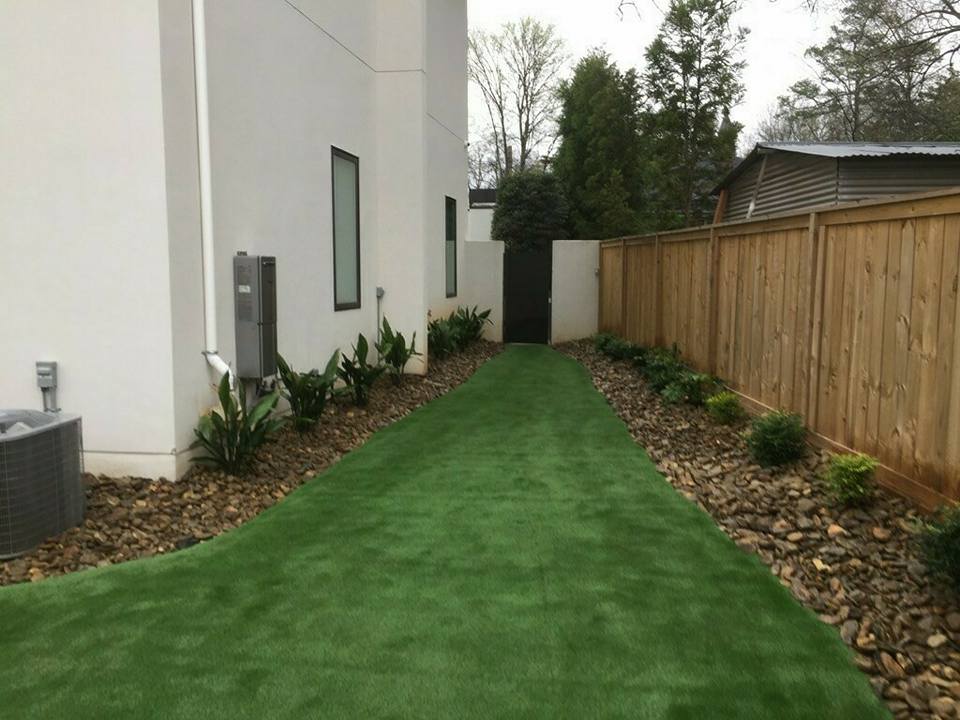 A narrow yard with artificial grass leads to a gated fence. The area is bordered by white walls on one side and a wooden fence on the other, with decorative pebbles and small plants lining the edges.