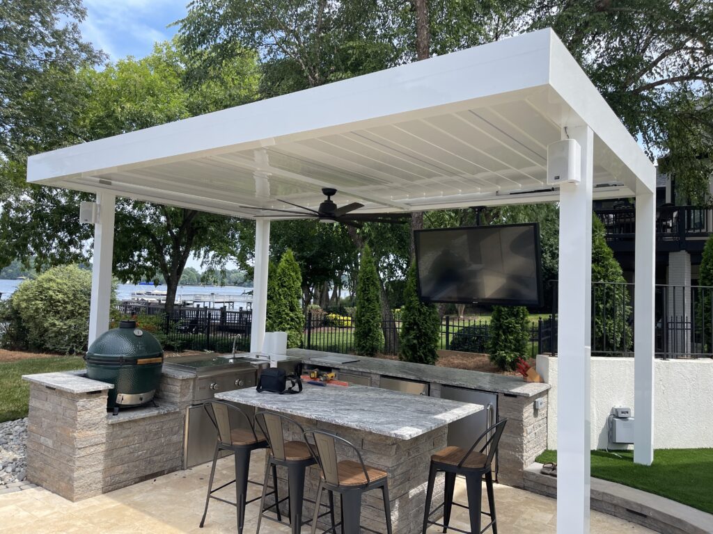 Outdoor kitchen with a white pergola, featuring a grill, sink, and marble countertop with barstools. A TV is mounted above the counter. Lush green trees and a view of the water are visible in the background.