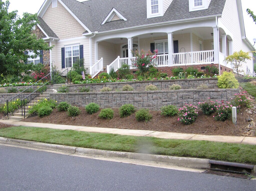 A suburban house with a well-maintained, multi-tiered garden. The garden includes various shrubs and flowers, bordered by a stone retaining wall. A staircase and a grassy lawn lead up to the house's porch.