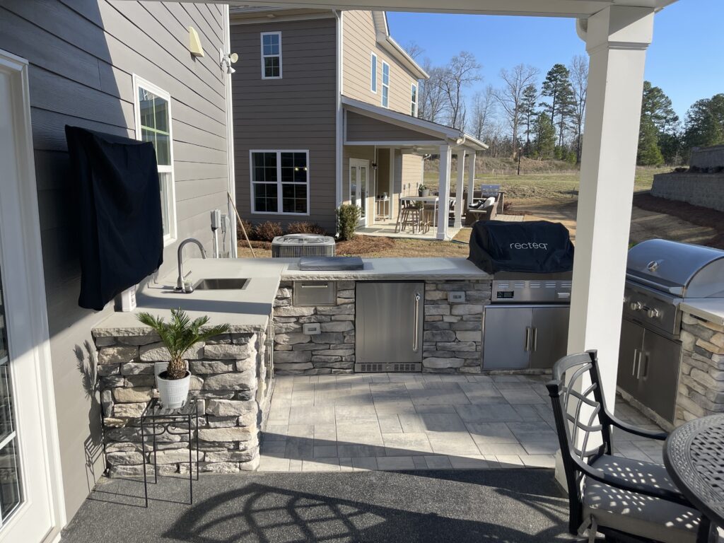 Outdoor kitchen with stone counters, a built-in grill, sink, and mini fridge. Covered grill with a "rectec" cover. Adjacent patio furniture includes a round table and chairs. Two-story house and lawn visible in the background.