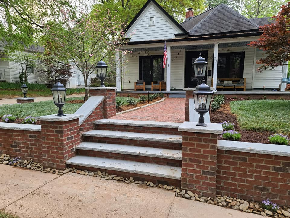 A charming house with a brick path and stairs leading to the front porch, adorned with four lantern-style lamps. The porch features two rocking chairs, and an American flag is displayed. Green lawns and shrubs surround the home.