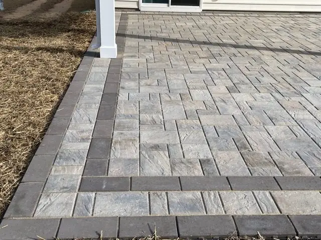 A gray brick patio with a herringbone pattern is bordered by darker bricks. The patio is adjacent to a building and surrounded by areas of bare soil.