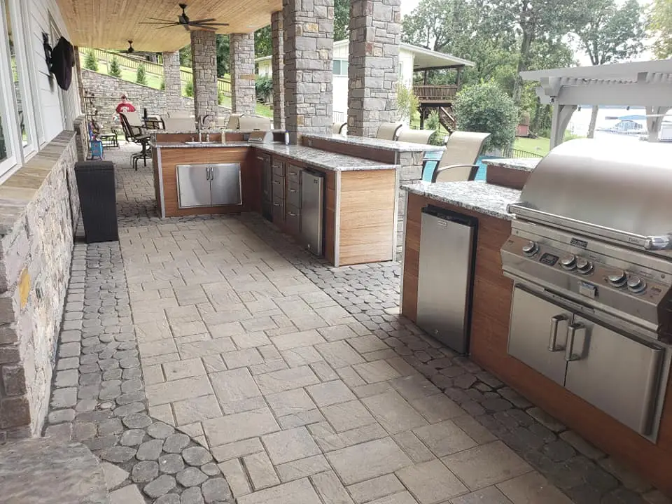Outdoor kitchen with stainless steel appliances, including a grill, refrigerator, and storage cabinets. Stone and wood accents complement the space, which is set on a patio with a stone-paved floor under a wooden ceiling. Outdoor seating is visible in the background.