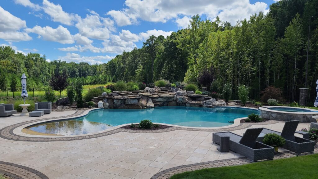 A luxurious outdoor pool with a waterfall feature and surrounding lounge chairs is set against a backdrop of lush green trees and a bright blue sky with scattered clouds. The patio is tiled and spacious, offering a serene and inviting atmosphere.