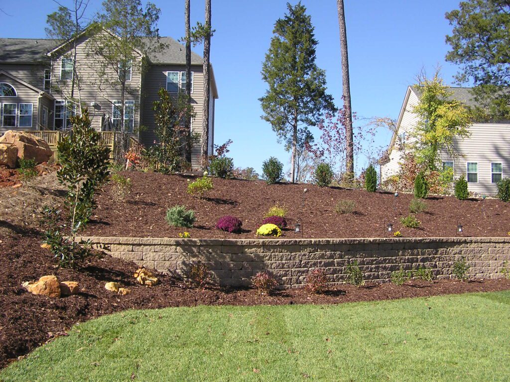 A landscaped garden on a slope with a stone retaining wall. The garden features various shrubs and plants, with two houses visible in the background and a clear blue sky above.