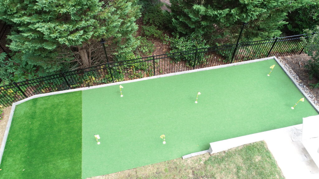 Aerial view of a rectangular mini-golf putting green surrounded by a black fence and lush trees. The green has five small flags indicating hole locations. Adjacent is a patch of slightly darker grass. Stone steps are visible on one side.