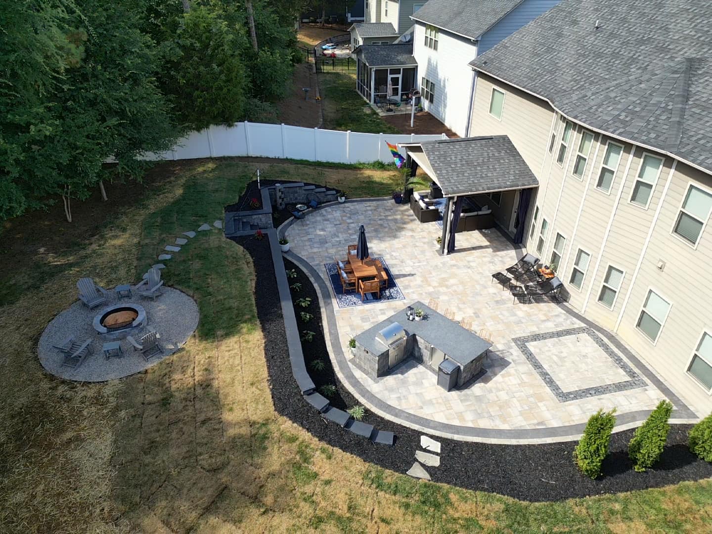 Aerial view of a backyard with a stone patio featuring a dining set, a covered outdoor kitchen, and a small bar area. A curved path leads to a circular fire pit with chairs. The yard is bordered by trees and a white fence.