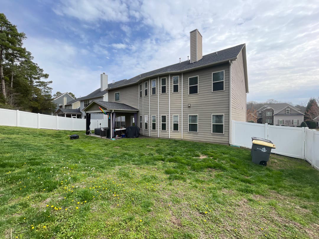 A large, two-story beige house with a chimney stands in a grassy backyard. It's enclosed by a white fence. The yard features a small playhouse, a barbecue grill, and a trash bin. The sky is partly cloudy.