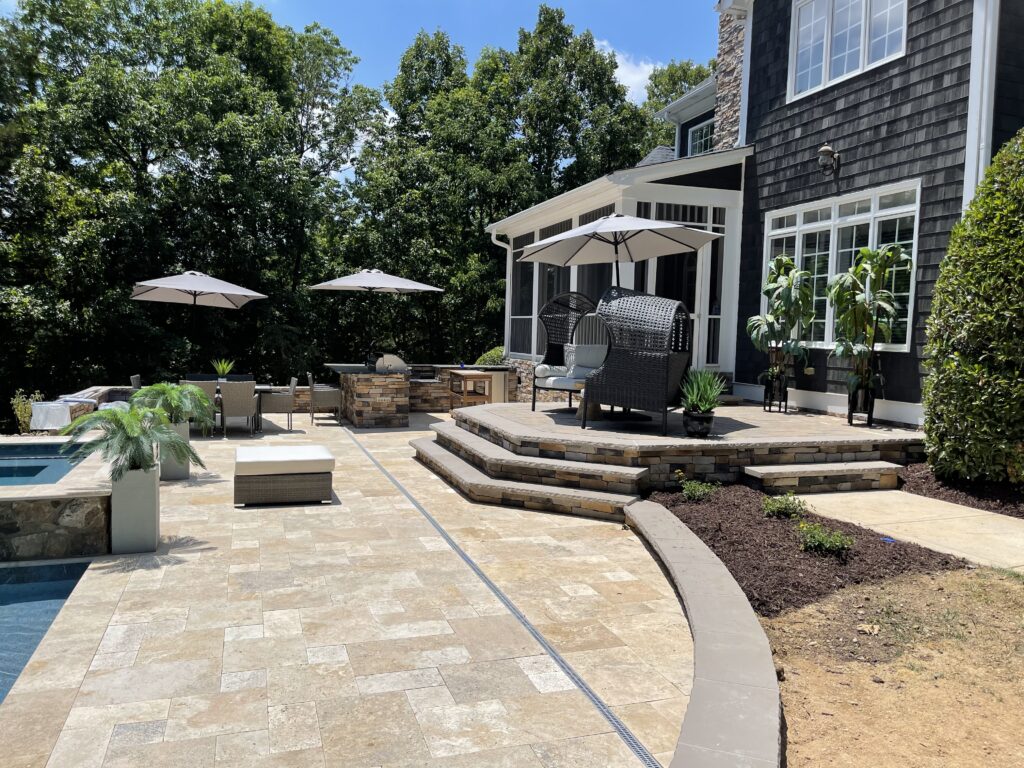 A modern outdoor patio with a stone-tiled floor features a pool, lounge chairs, umbrellas, and a seating area with tables. The multi-tiered design includes stairs leading to a covered porch attached to a house, surrounded by lush greenery.