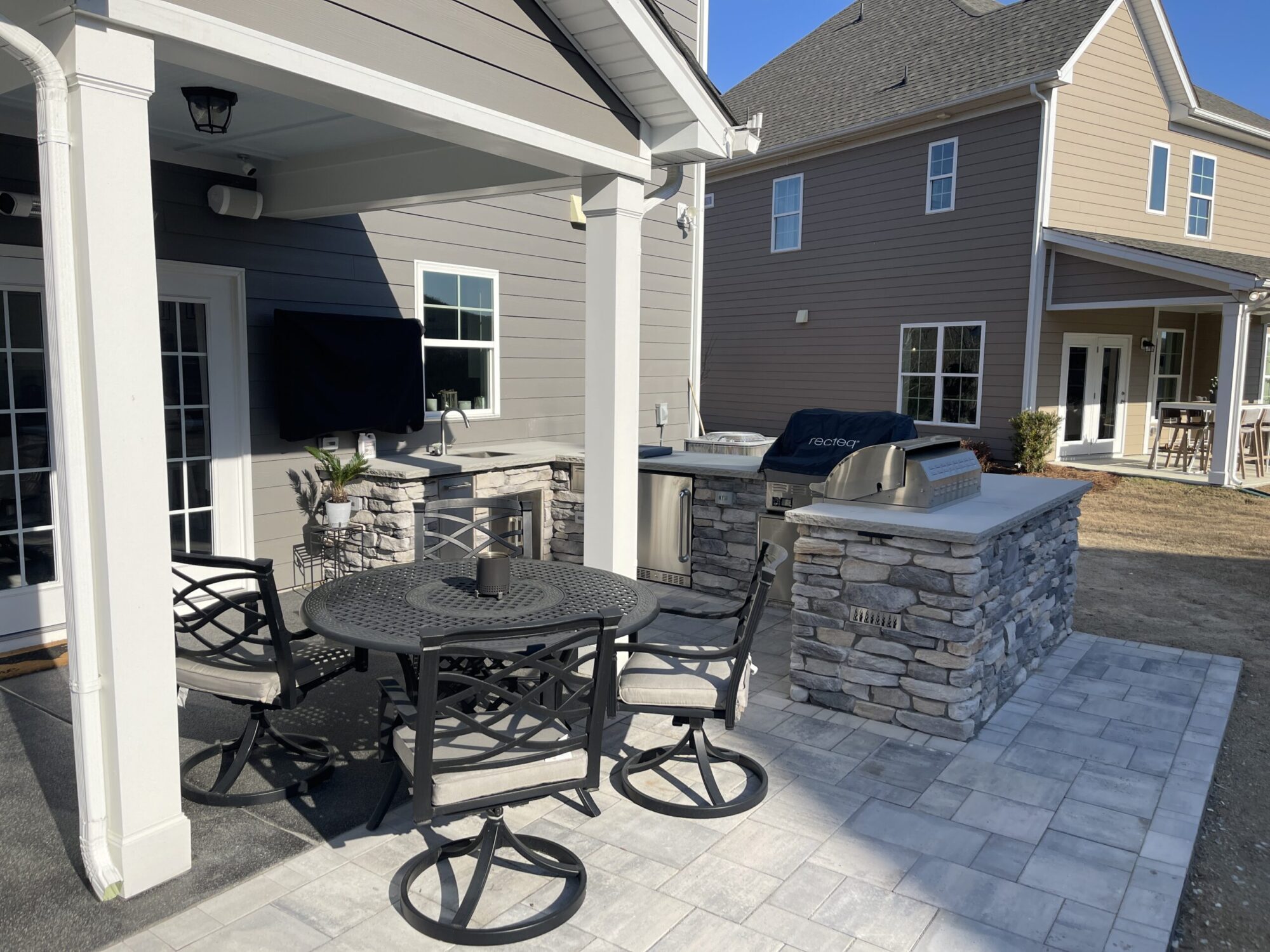 Outdoor patio with a round metal table and four chairs. There's a built-in grill and countertop with stone finish, shaded by a roof, next to a TV on the wall. The patio is part of a modern house with another house visible in the background.